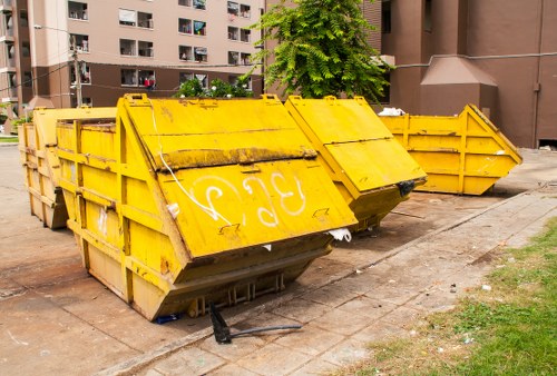 A row of large, heavy-duty industrial skip containers painted bright yellow, positioned on a paved area next to a patch of grass and in front of a multi-storey residential building. The skips are constructed from metal with a ribbed texture and have high, reinforced sides, some displaying faded graffiti or markings on their surfaces. The lids on top of the skips are partially open or tilted, revealing dark interiors. The skips are aligned in a slightly staggered manner, with some placed directly on the pavement and others slightly elevated on the edges. Surrounding environment includes a concrete sidewalk, a small grass strip with a few patches of soil, and nearby trees offering partial shade. The background features a brown brick building with multiple balconies and windows. This setup is typical for waste removal or clearance services in Shepherd's Bush, and the company Shepherds Bush House Clearance may use such containers for rubbish collection or clearance procedures.