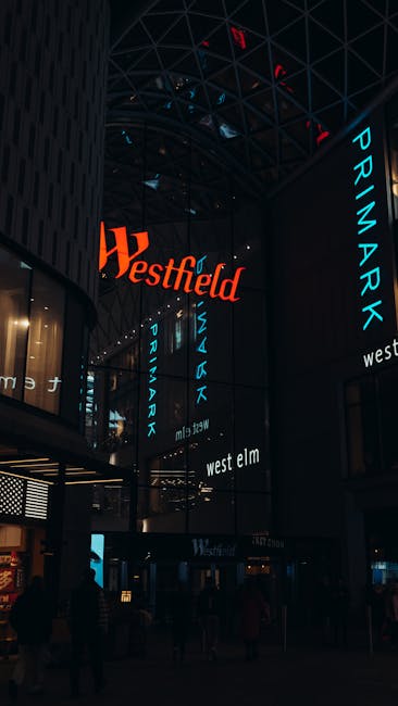 The image depicts a modern, multi-storey commercial building at night, featuring large illuminated signs displaying the words 'Westfield' in red and 'PRIMARK' in blue on the exterior facade. The build