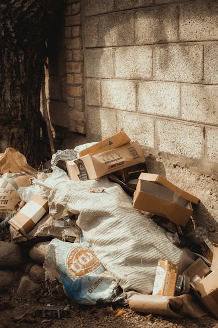 A pile of mixed waste materials is situated at the base of a concrete brick wall outdoors. The waste includes flattened cardboard boxes, some partially torn, and plastic bags filled with rubbish. The 