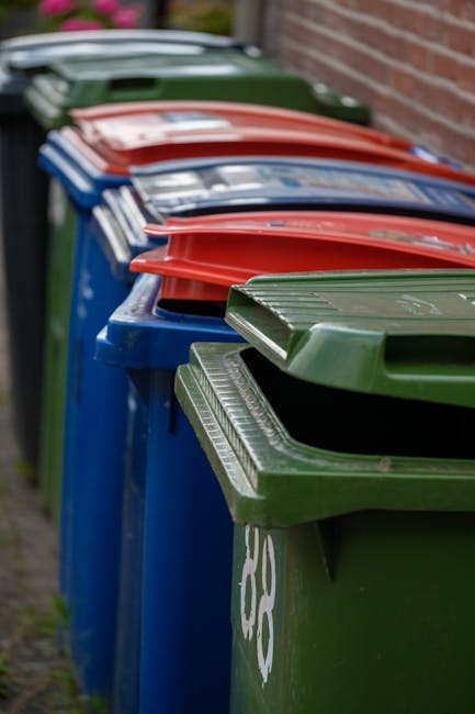 A pile of mixed waste materials is situated at the base of a concrete brick wall outdoors. The waste includes flattened cardboard boxes, some partially torn, and plastic bags filled with rubbish. The 